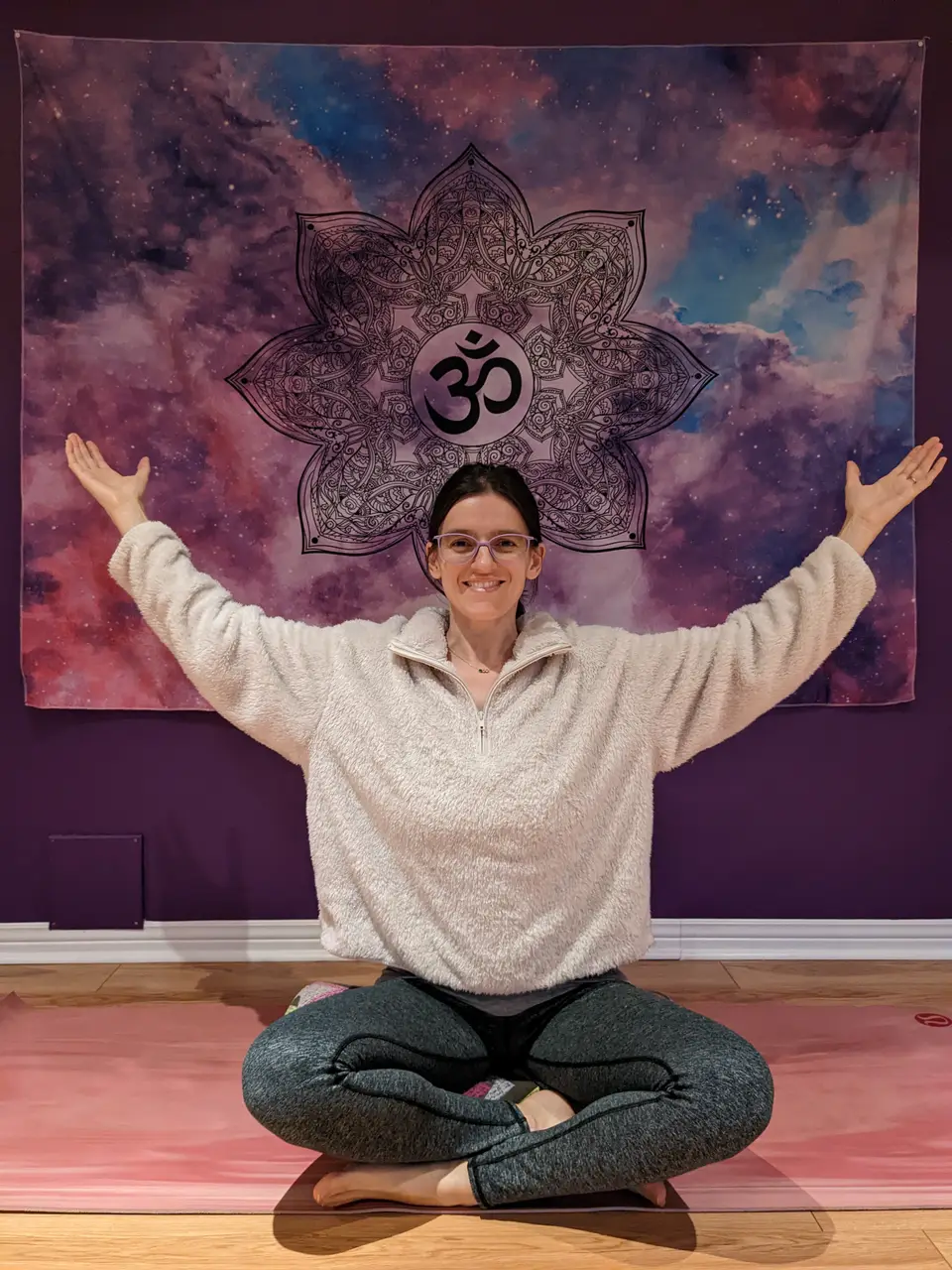 Jessica assise en lotus, bras ouverts, devant une tenture mandala violette